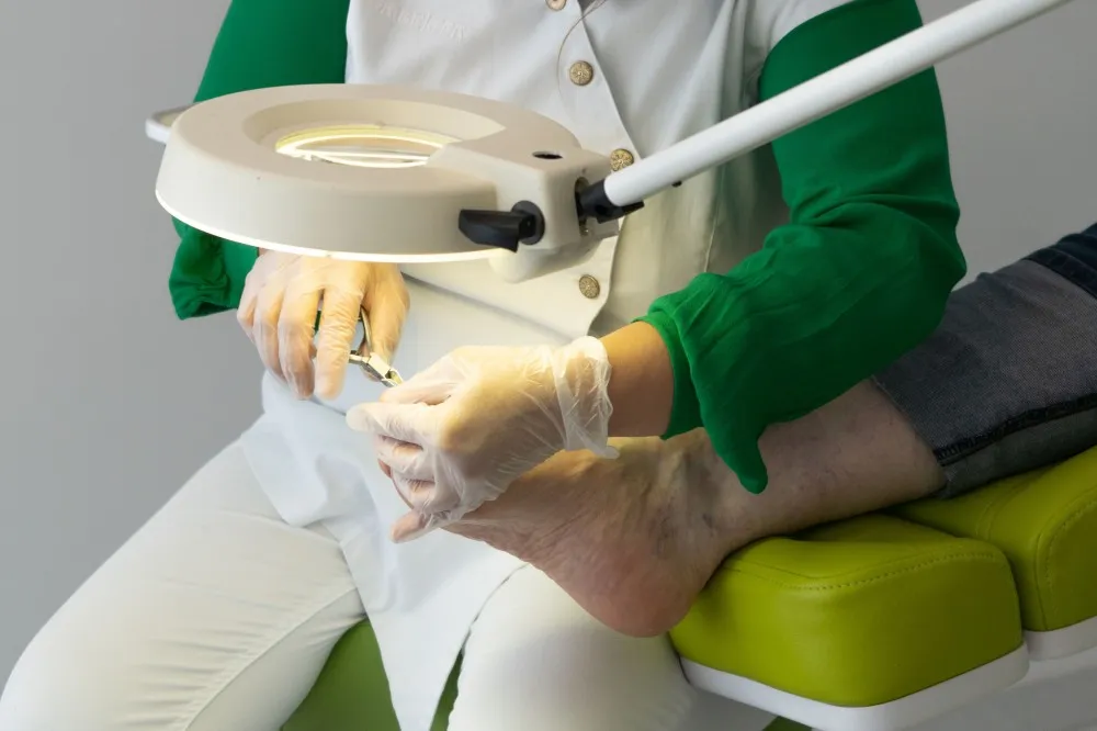 Close-up of a professional medical pedicure procedure being performed in a comfortable home setting. A therapist with soft-focus hands is carefully caring for a client's feet using high-end, clean equipment. The lighting is bright and clean but warm, emphasizing hygiene and expert care. The surrounding environment is a minimalist, luxury home with soft textures in cream and taupe.